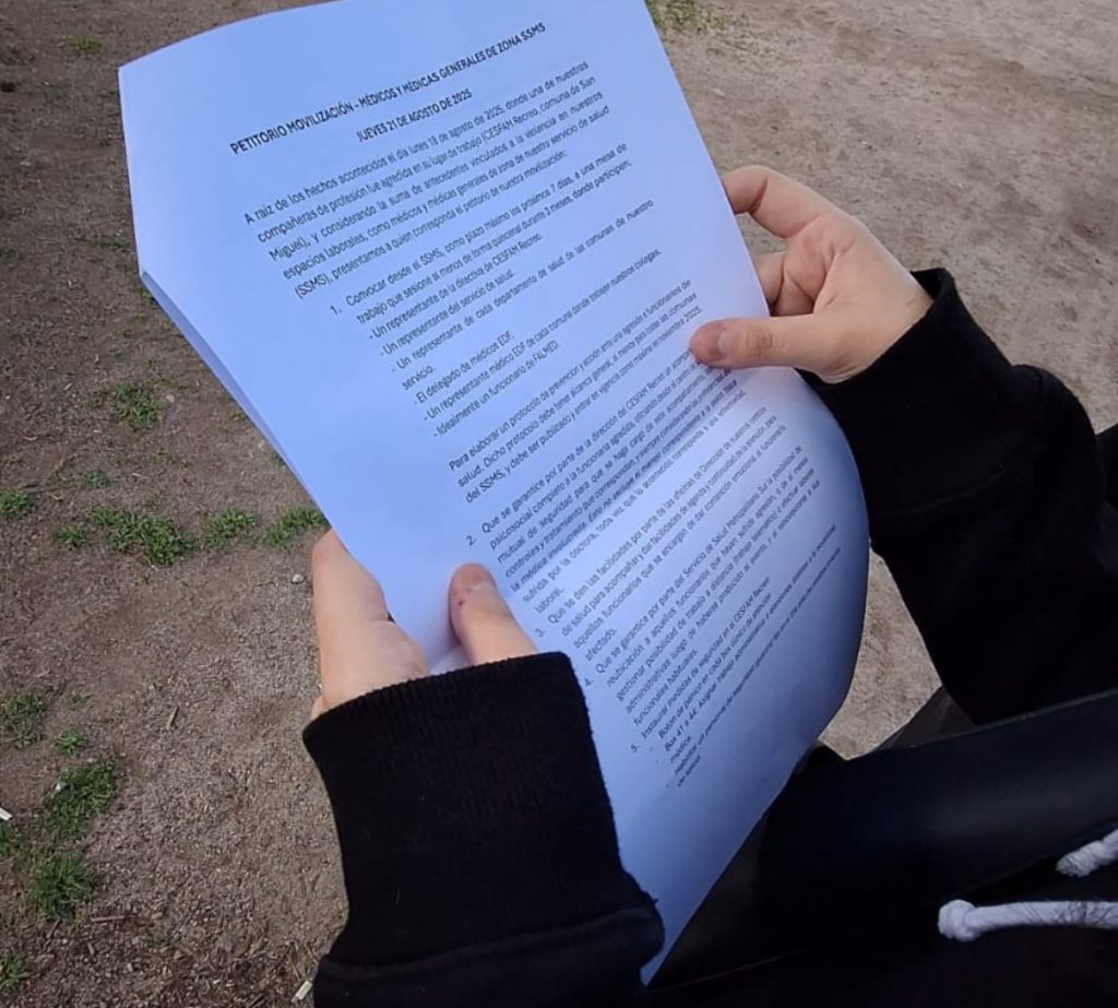 Fotografía de una hoja de papel con un petitorio en la descripción. Se ven dos manos y una chaqueta negra sujetando la hoja de papel. Atrás de la hoja hay un piso de tierra. 