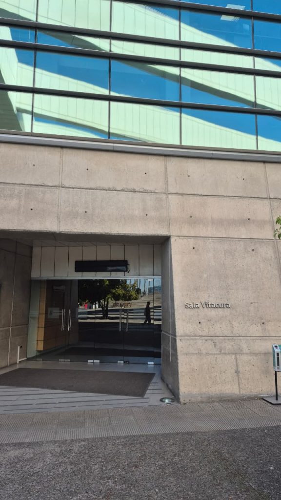 Se muestra la entrada principal de la “Sala Vitacura”, parte del Teatro Mori. Puerta cerrada y se refleja la silueta de una persona pasando. A la derecha de la puerta, la pared de hormigón exhibe discretamente el texto “sala Vitacura” grabado. Una alfombra oscura se extiende desde la entrada hacia el exterior.