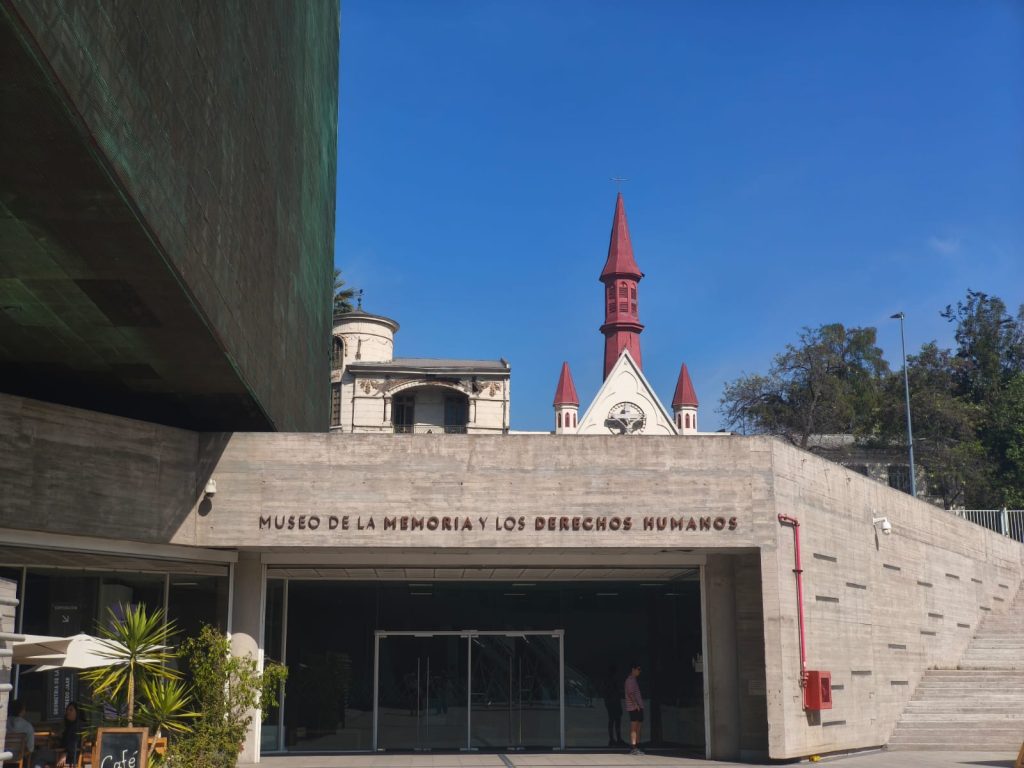 Esta imagen captura la entrada principal del Museo de la Memoria y los Derechos Humanos. A la izquierda, se ve parte de la fachada moderna del museo, mientras que en el centro y fondo, se eleva la torre del campanario de color rojo del Santuario Cristo Pobre, creando un contraste visual entre la arquitectura contemporánea del museo y el patrimonio histórico. El cielo es azul y despejado. Se pueden distinguir algunas personas en la entrada y un pequeño café y un área de descanso con plantas a la izquierda.