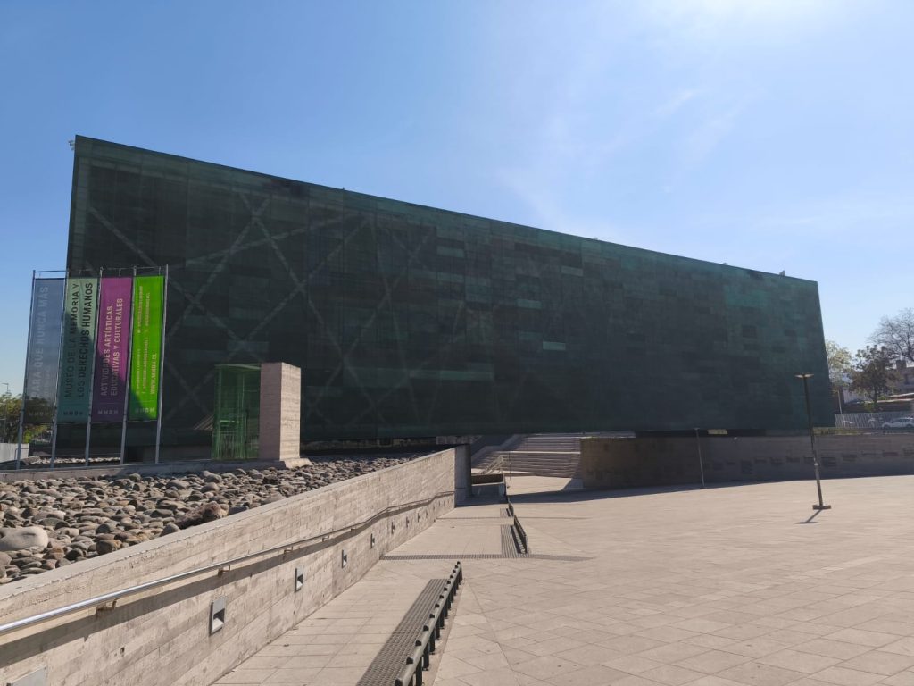 Fachada principal del Museo de la Memoria y los Derechos Humanos. El edificio es una estructura moderna, dominada por una gran superficie de paneles de color verde oscuro que reflejan el cielo. Frente a la entrada, se observan varias banderas verticales de colores (gris, morado y verde) con texto informativo. La escena se desarrolla en una plaza pública pavimentada con baldosas claras, donde también se aprecia una pared baja de piedra. El cielo es completamente azul y despejado, indicando un día soleado.
