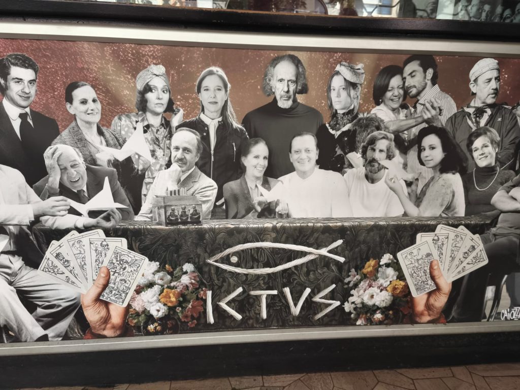 Fotografía artística en blanco y negro que muestra a un elenco de teatro reunido en una composición similar a "La Última Cena". La imagen, que parece un cartel, mezcla el blanco y negro del grupo con el color de unas manos sosteniendo cartas del tarot en la parte inferior. En el centro de la mesa se destaca el logo del pez y el nombre "ICTUS", asociado al Teatro ICTUS.